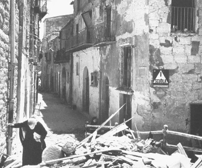 Sicilia, 1943. Un'anziana donna tra le rovine del suo paese. Sicily, 1943. And old woman walking in the ruins of her village.